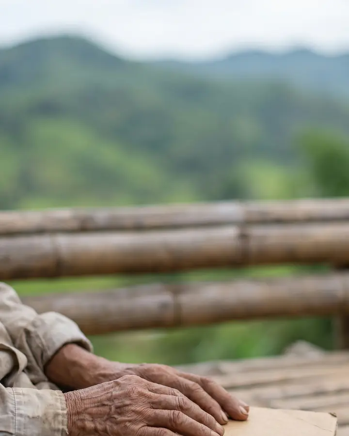 An older person's hands resting on a closed plain folder on a bamboo bench, with green hills behind.