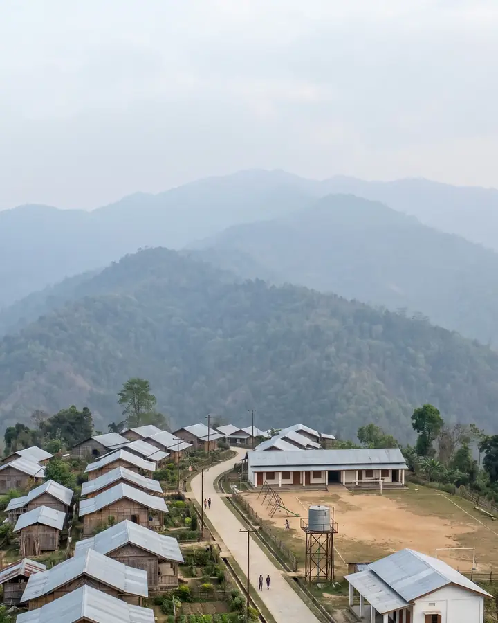 A planned hill village with tin-roof houses and a road, mostly covered by a grid of 25 icon cards with large labels. Cards: Roads, Drainage, Drinking water, Water for cattle, Grazing land, Fair price shop, Panchayat Bhawan, Post office, Seed & fertiliser store, Irrigation, Public transport, Burial / cremation, Toilets & sanitation, Electricity, Anganwadi, School, Sub-health centre (2 km), Health centre, Playground, Community centre, Places of worship, Land for traditional institutions, Forest produce rights, Security, Veterinary centre.